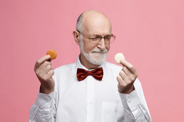 Еда, сладости и концепция угощений. нерешительный старший бородатый мужчина, сладкоежка, держит два разноцветных печенья macarons, хмурится, выбирает между ними, носит очки и галстук-бабочку