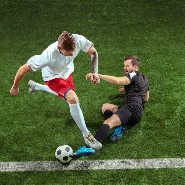 Football player tackling for ball over green grass wall. Professional male soccer players in motion at stadium. Fit jumping men in action, jump, movement at game.