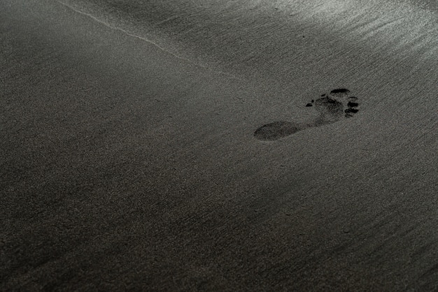 黒い砂のビーチのマクロ写真の足跡。フィールドの浅い深さで絹のような黒いビーチテクスチャの人間の跡。ミニマルな黒の背景。テネリフェ島のヴォルカニック砂浜。