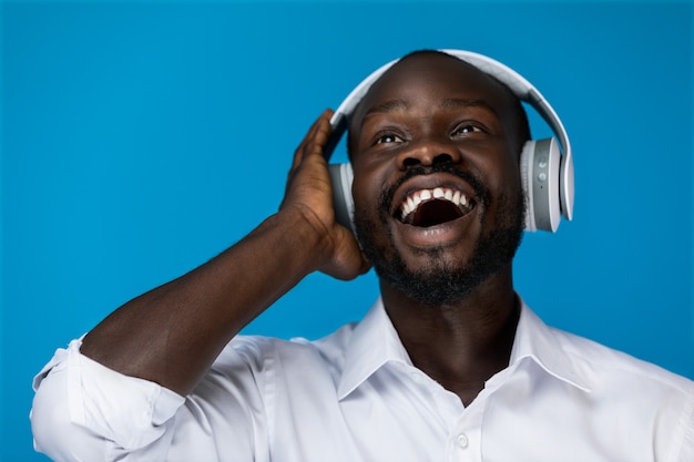 Foreground bearded smiling afroamerican man with open eyes looking up is holding by one hand big headphones