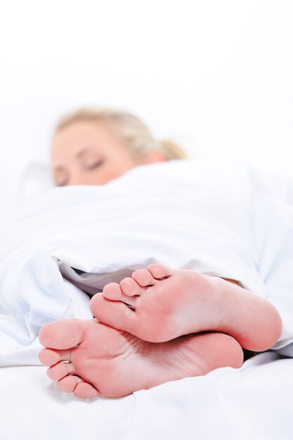 На переднем плане крупным планом чистые ноги спящей симпатичной женщины прикрывают белое одеяло