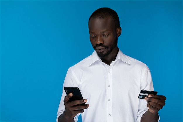 Foreground thoughtful bearded afro american guy is looking at cell phone and holding credit card