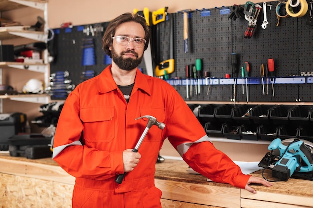 Мастер в оранжевой рабочей одежде и защитных очках держит молоток, мечтательно глядя в камеру с инструментами на заднем плане в мастерской