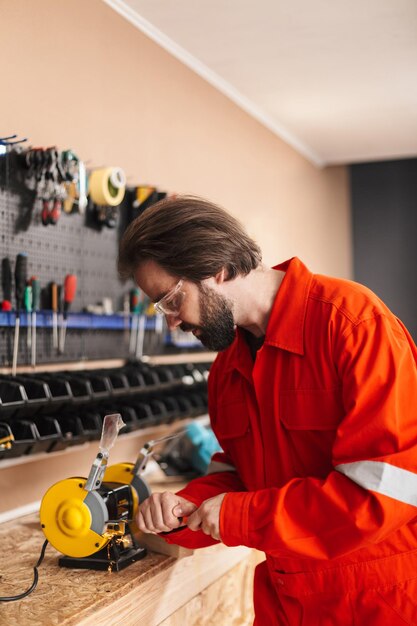 Foreman in orange work clothes and protective eyewear thoughtfully working with knife sharpener in workshop