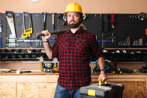 Foreman in yellow hardhat holding hammer on shoulder and toolbox in hand dreamily looking in camera in workshop