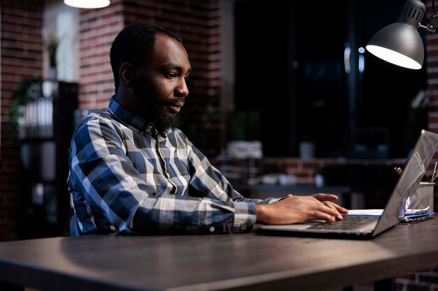 Forex stock market professional trading agent following real time financial graphs and investment profits. African american company investor sitting at desk in office workspace at night.