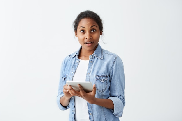 Forgetful Afro-American female with dark appealing in blue shirt holding tablet in her hands staring  with astonished amazed look, having completely forgotten about something serious.