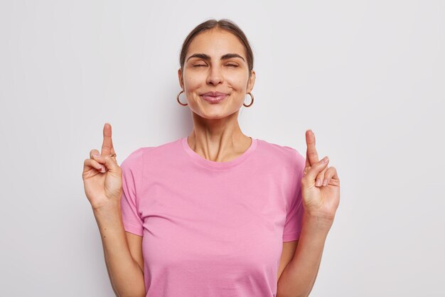 Fortune please come. Pleased brunette woman keeps fingers crossed makes wish prays and hopes for miracle anticipates for something desirable wears pink t shirt isolated over white background