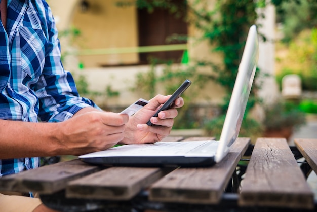 Forty years old caucasian man looking at credit card while working on laptop computer on garden terrace during sunny summer day. Modern lifestyle - countryside weekend and shopping online concept.