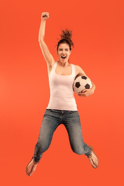 Free photo forward to the victory.the young woman as soccer football player jumping and kicking the ball at studio on a red background. football fan and world championship concept. human emotions concepts