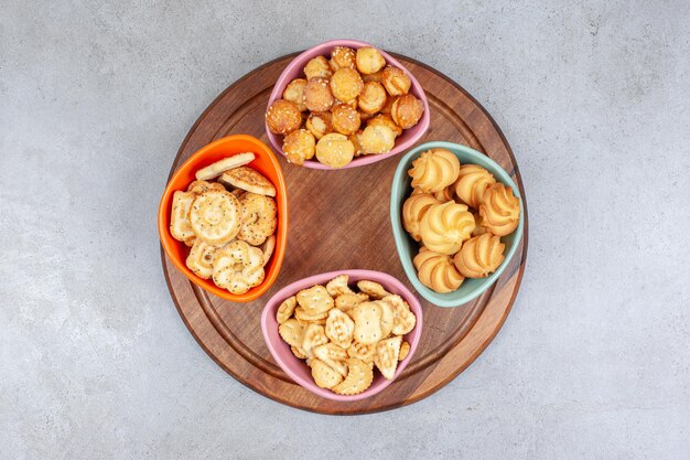 Four bowls of biscuit and cookie chips on wooden board on marble surface.