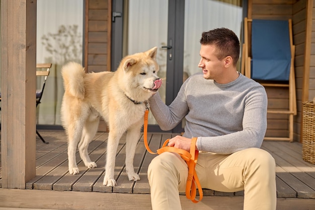 4本足の友達。晴れた日にひもで生姜犬に触れて外を見ているカントリーハウスのポーチに座っているカジュアルな服を着た若い大人の笑顔の男