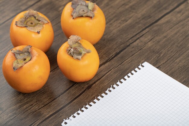 Four ripe persimmon fruits and empty notebook on wooden surface