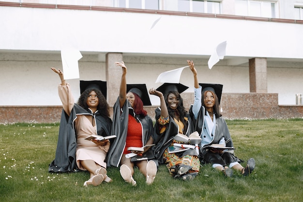 Free photo four young afro american female students dressed in black graduation gown. campus as a background