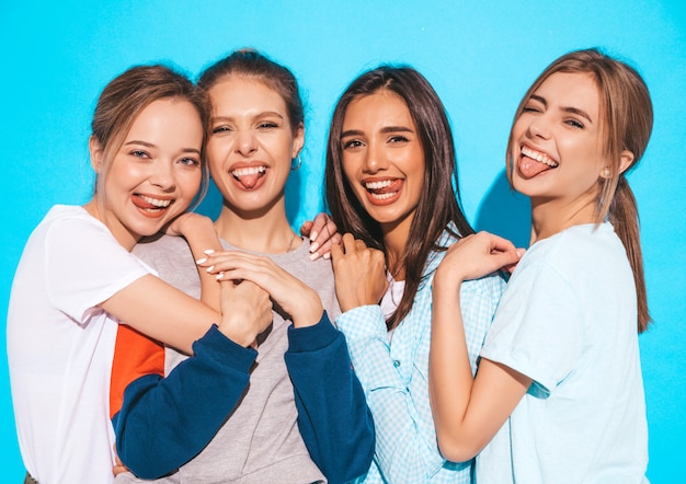 Four young beautiful smiling hipster girls in trendy summer clothes. Sexy carefree women posing near blue wall in studio. Positive models having fun and hugging.They show tongues