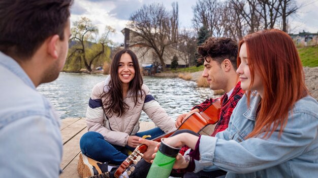 Four young friends singing, resting and playing guitar near a lake in a park