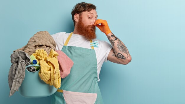 Foxy bearded man closes nose with fingers from unpleasant smell, collects all dirty laundry, wears casual t shirt and apron with clothespins, has tattoo, stands over blue wall, free space