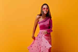 Free photo frail girl in stylish pleated skirt and heart-shaped glasses moves her outfit. stylish portrait in full growth on orange wall