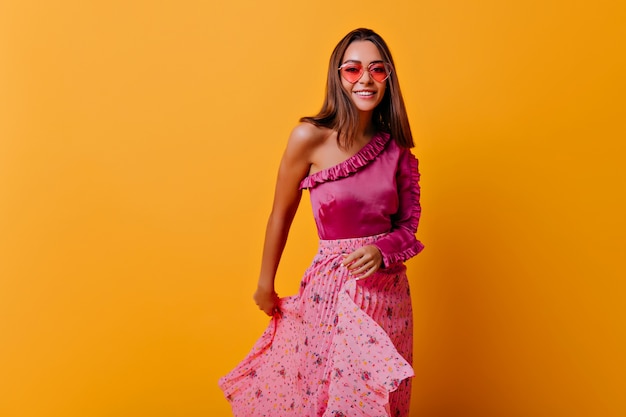 Frail girl in stylish pleated skirt and heart-shaped glasses moves her outfit. Stylish portrait in full growth on orange wall