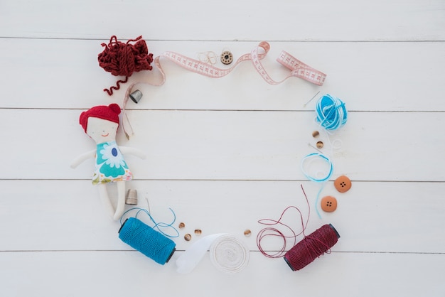 A frame made with rag doll; thimble; wool; measuring tape; button; ribbon and yarn spool on wooden desk