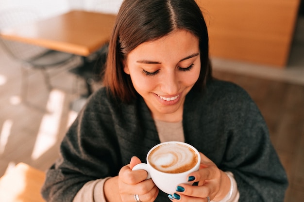 Кадр выше улыбающейся счастливой темноволосой женщины, пьющей кофе в кафе