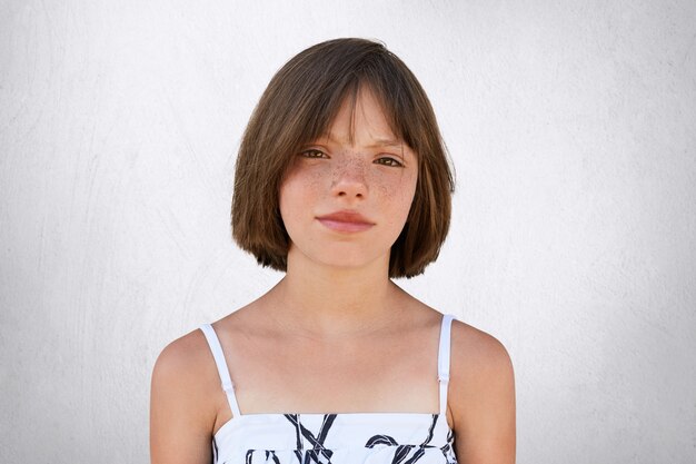 Freckled girl with hazel eyes and dark short hair, looking with displeasure into camera while posing on whiye concrete wall. Beautiful little girl with stylish hairdo wearing summer dress