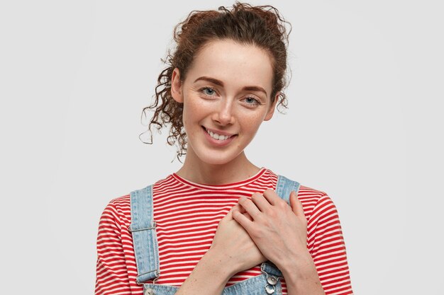 Freckled young curly female keeps hands on heart, expresses good attitude, dressed casually, shows natural beauty, being in high spirt, isolated over white wall. Friendly smiling girl.