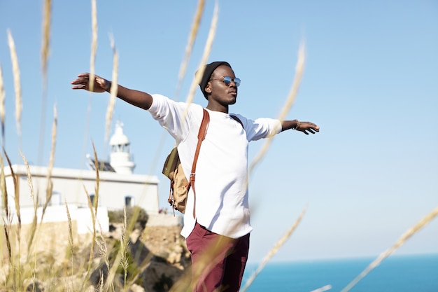 Free happy stylish male tourist having relaxed and carefree look while standing on edge of cliff, spreading arms like a bird, feeling warm wind on sunny day during his journey abroad. Summer concept