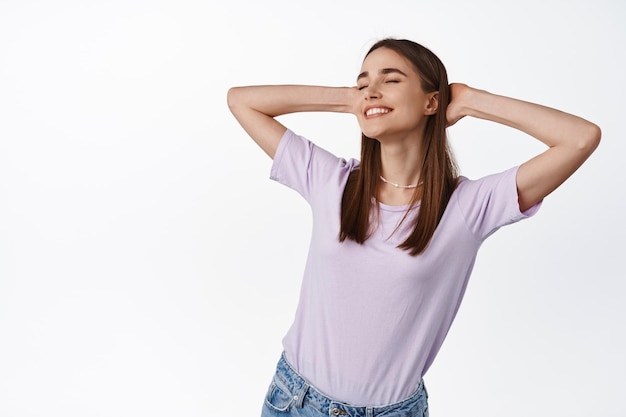 Free and relaxed young woman relaxing, close eyes and smile, holds hands behind head in resting pose, as if lying under sun on summer holiday, vacation concept, white background