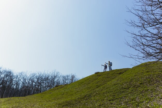 Freedom all the time. Aged family couple of man and woman in tourist outfit walking at green lawn near by trees in sunny day. Concept of tourism, healthy lifestyle, relaxation and togetherness.