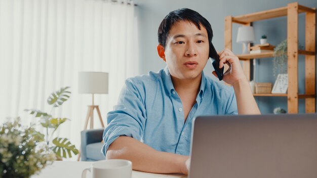 Freelance Asia guy casual wear using laptop talk on cell phone in living room at house.