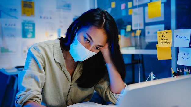 Freelance asia women wear face mask using laptop hard work at new normal office. working from home overload at night, self isolation, social distancing, quarantine for corona virus prevention.