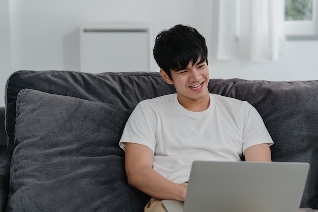 Freelance Asian man working at home, male creative on laptop on sofa in living room. Business young man owner entrepreneur, play computer, checking social media in workplace at modern house .