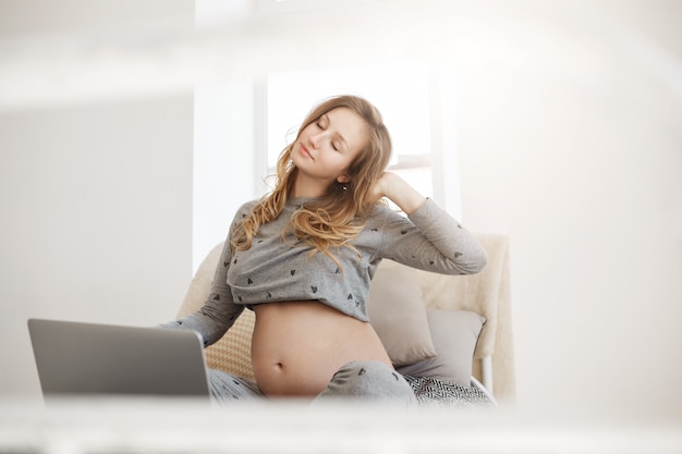 フリーランス、ビジネスコンセプト。ラップトップコンピューターでの長時間の作業の後にストレッチして、寝室に座っている灰色のホームウェアの若い成功した妊娠中の女性フリーランスデザイナーの肖像画。