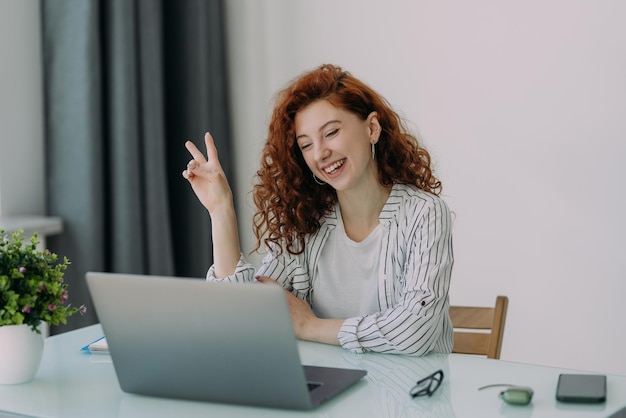 Freelance business woman with ginger hair sits in front of laptop computer communicates with colleagues via video conference sits at desktop drinks coffee has happy expression Distance job