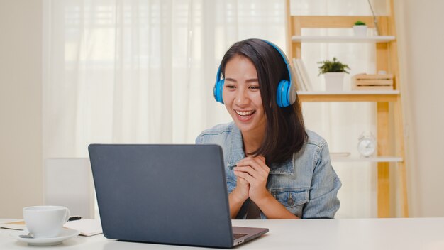 Freelance business women casual wear using laptop working call video conference with customer in workplace in living room at home. Happy young Asian girl relax sitting on desk do job in internet.