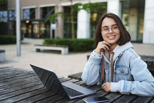 Concetto di carriera e gioventù freelance felice giovane donna spensierata seduta all'aperto su una panchina che lavora con laptop e smartphone guardati intorno sorridendo prendi ispirazione mentre scrivi un saggio