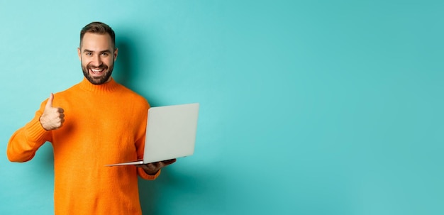 Foto gratuita freelance e concetto di tecnologia uomo fortunato in maglione arancione che mostra il pollice in su mentre si lavora con il giro