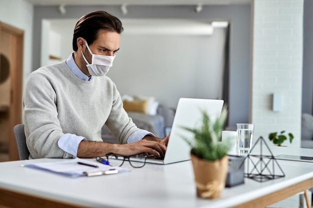 Freelance worker using laptop and wearing protective face mask while working at home during coronavirus epidemic