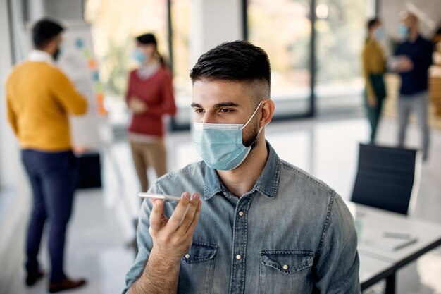 Freelance worker with face mask talking over cell phone's speaker in the office