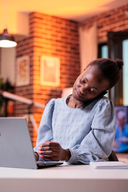 Free photo freelancer answering phone call to discuss work plan with clients in modern home office with beautiful warm sunset light. african american woman chatting with company colleagues on smartphone