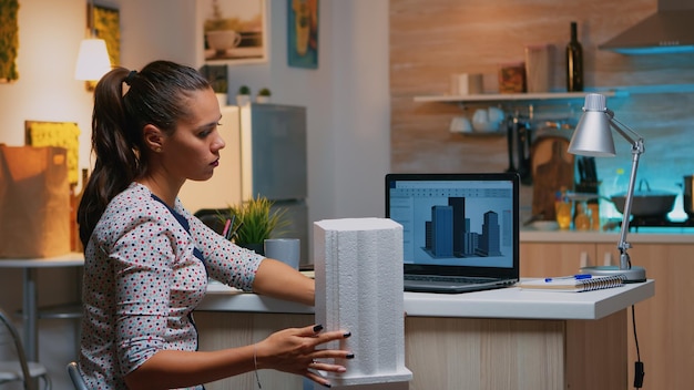 Free photo freelancer architect working in 3d software to elaborate buildings design sitting at kitchen desk at night. engineer artist creating and studying in office holding scale model, determination, career.