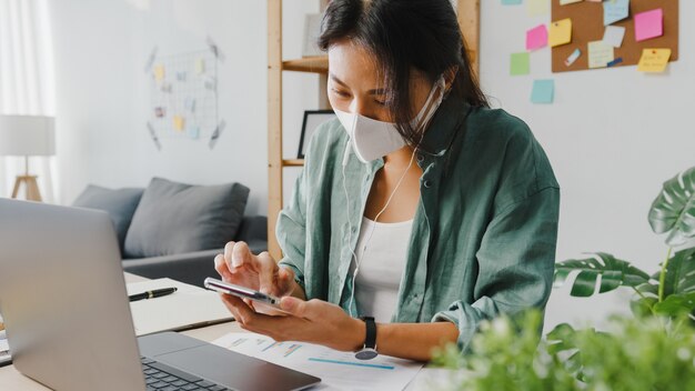 Freelancer Asia women wear face mask using smartphone shopping online via website while sitting at desk in living room.