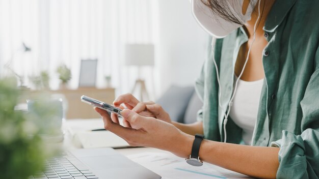 Freelancer Asia women wear face mask using smartphone shopping online via website while sitting at desk in living room.