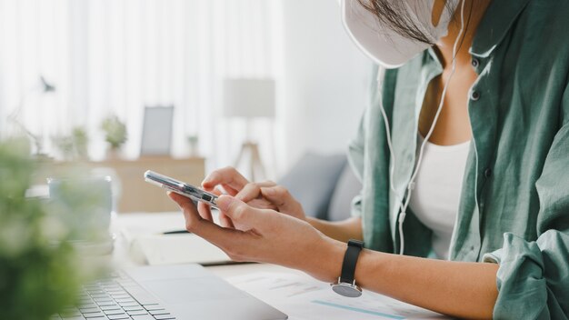 フリーランサーアジアの女性は、リビングルームの机に座っている間、ウェブサイトを介してオンラインショッピングのスマートフォンを使用してフェイスマスクを着用します。