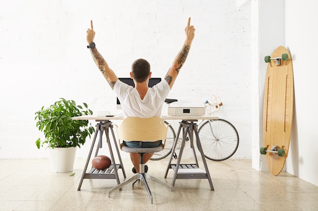 Free photo freelancer at his desktop with personal computer, surrounded with his hobby toys in light room in front of white brick wall, extends his arms and shows uncensored gesture