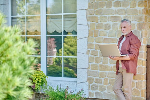 Freelancer. Mature man in casual clothes with a laptop near a brick wall