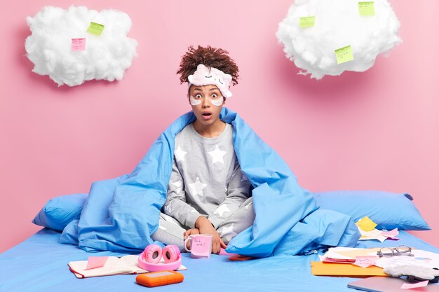 freelancer sits in lotus pose on comfortable bed stares impressed wears pajama prepares report surrounded by papers and sticky notes isolated on pink