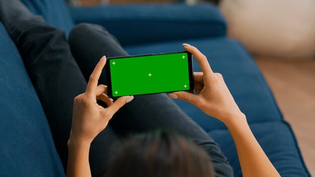 Фрилансер сидит на диване и смотрит фильмы по телефону в горизонтальном режиме с макетом зеленого экрана с цветными клавишами. Женщина, использующая изолированное сенсорное устройство для просмотра социальных сетей