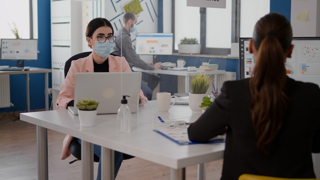 コロナウイルスの感染を防ぐために保護フェイスマスクを着用して新しい通常のオフィスに座っている間、ビジネスの開始について同僚と話しているフリーランサー。社会的距離を尊重するチーム